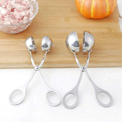 Close-up of two silver meatball makers, a bowl of raw ground meat, and an orange pumpkin on a wooden cutting board and white surface.