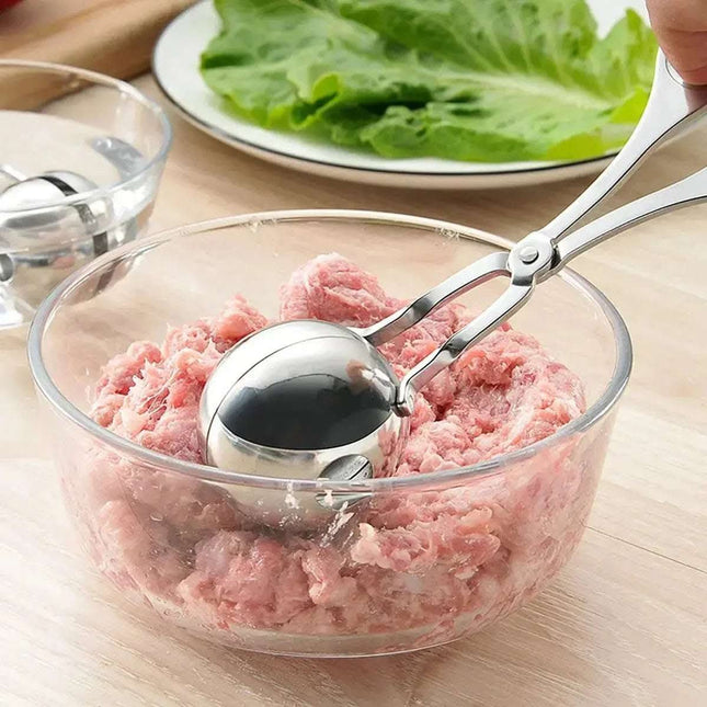 Close-up of a metal meatball maker scooping raw ground meat from a glass bowl, with a bowl of meatballs and a plate of lettuce in the background.